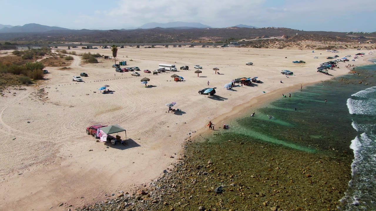 Wide drone view of beachgoers, cars, umbrellas, and green-blue surf