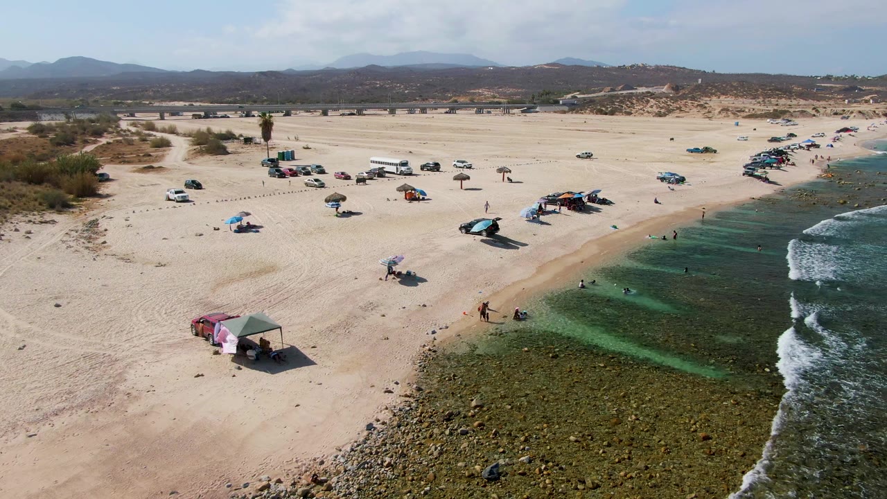 El Tule Beach Los Cabos appears from above with small figures in the water and on the sand, several umbrellas and vehicles scattered across the beach, and low hills beyond the dunes.