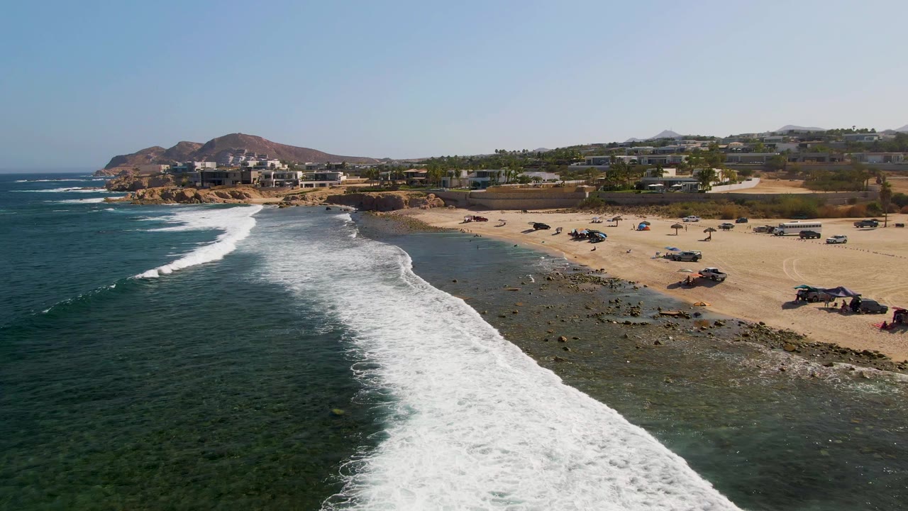 At El Tule Beach in Los Cabos, white surf approaches a rocky sandy shoreline with parked vehicles and hillside buildings in the distance, with no visible people.