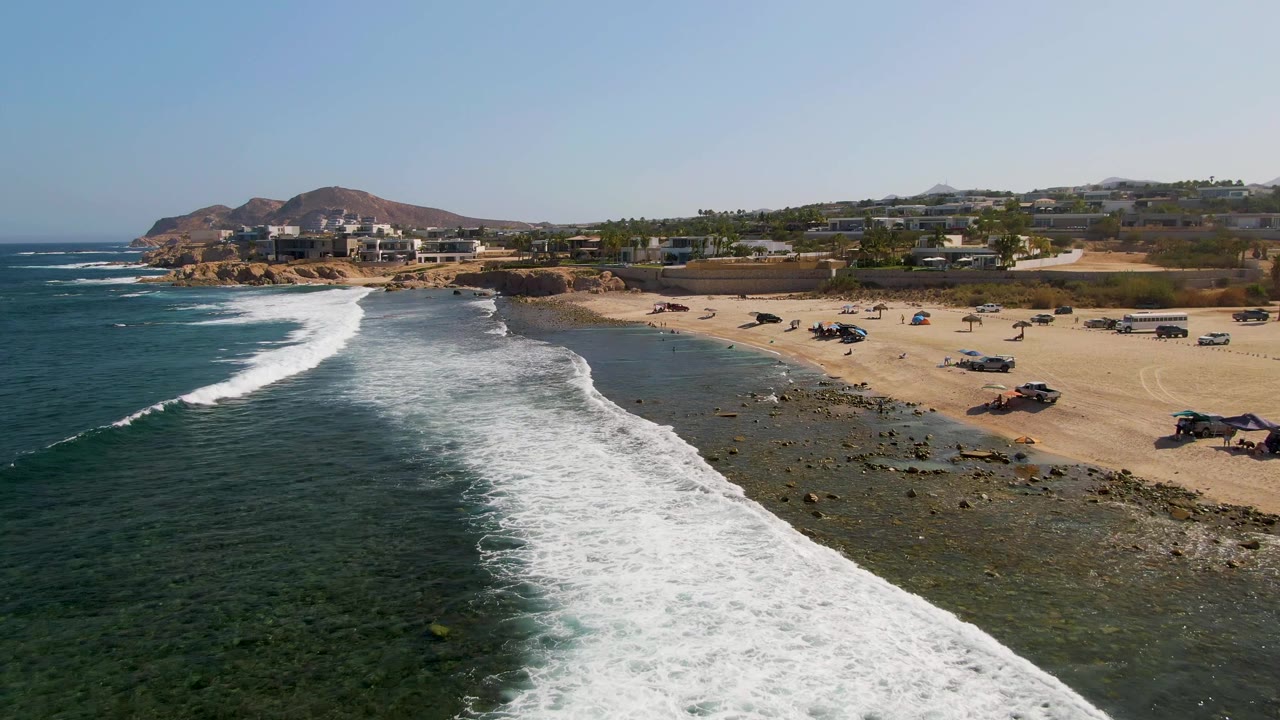 White wave line curves toward a beach with vehicles parked on sand.