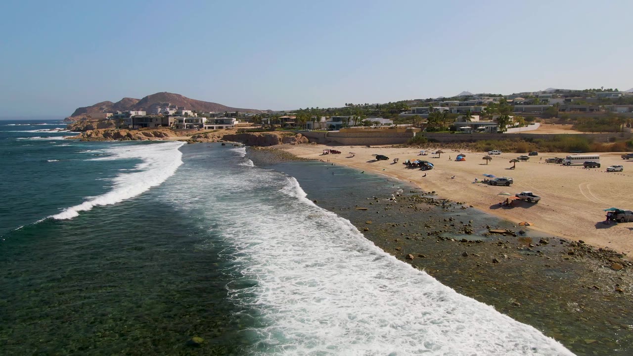 At El Tule Beach in Los Cabos, foamy water spreads wider over the shallow rocky edge as parked vehicles line the sand and low buildings remain in the background, with no visible people.