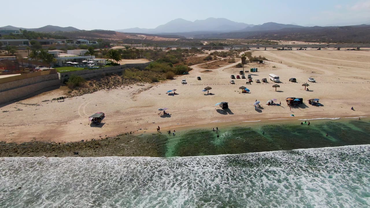 Aerial frame at El Tule Beach in Los Cabos showing foamy surf in the foreground, scattered umbrellas and small visible people on the sand, and dry hills beyond the beach.