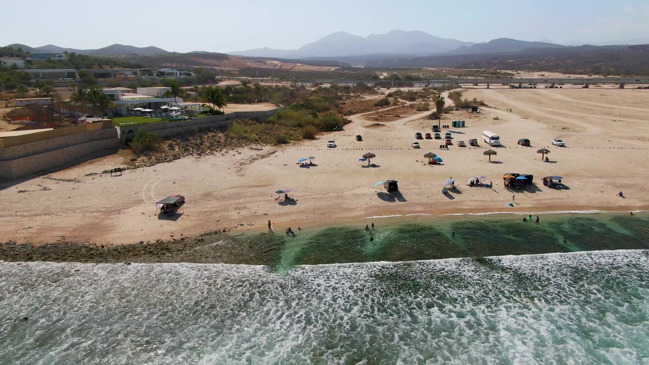 Waves roll toward a sandy beach with umbrellas, cars, and mountains in the distance.