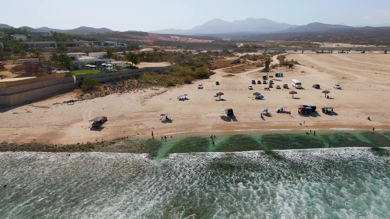 El Tule Beach appears from a high aerial angle with textured surf across the foreground, scattered visitors near the waterline, umbrellas on the sand, and low hills in the background.