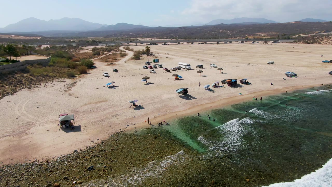 Wide aerial frame of El Tule Beach Los Cabos with rocky shallow water in the foreground, scattered cars and umbrellas on the sand, small visible people near shore, and mountains in the distance.