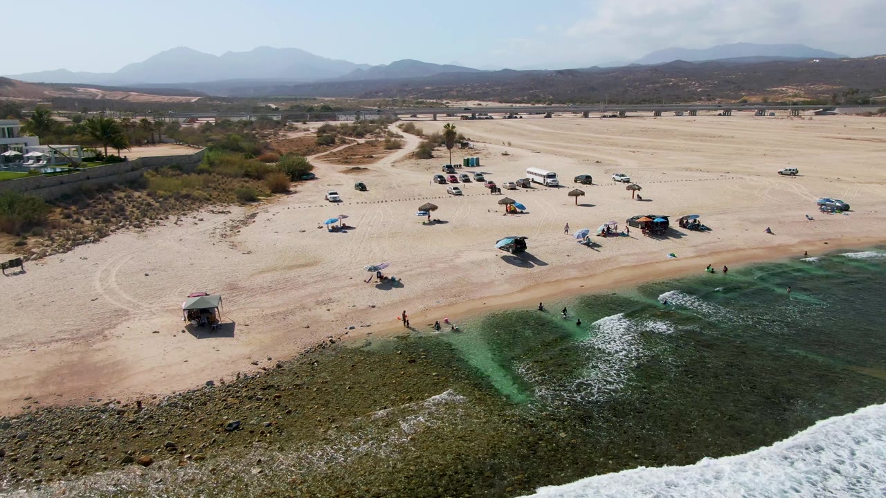 Aerial beach view with shallow green water, foam, parked vehicles, umbrellas, and distant mountains.