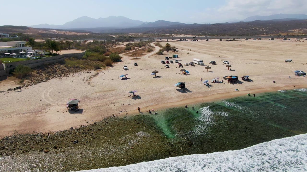 Aerial view over El Tule Beach Los Cabos as a brighter band of foam reaches the rocky foreground, with scattered beachgoers, cars, shade umbrellas, and desert hills behind the beach.