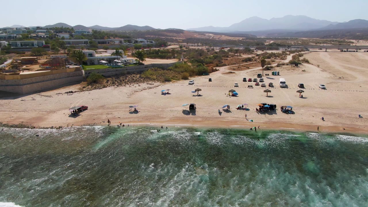 Aerial view at El Tule Beach in Los Cabos with green-blue surf in the foreground, a sandy beach with parked vehicles and umbrellas, scattered people, low buildings, dry hills, and distant mountains.