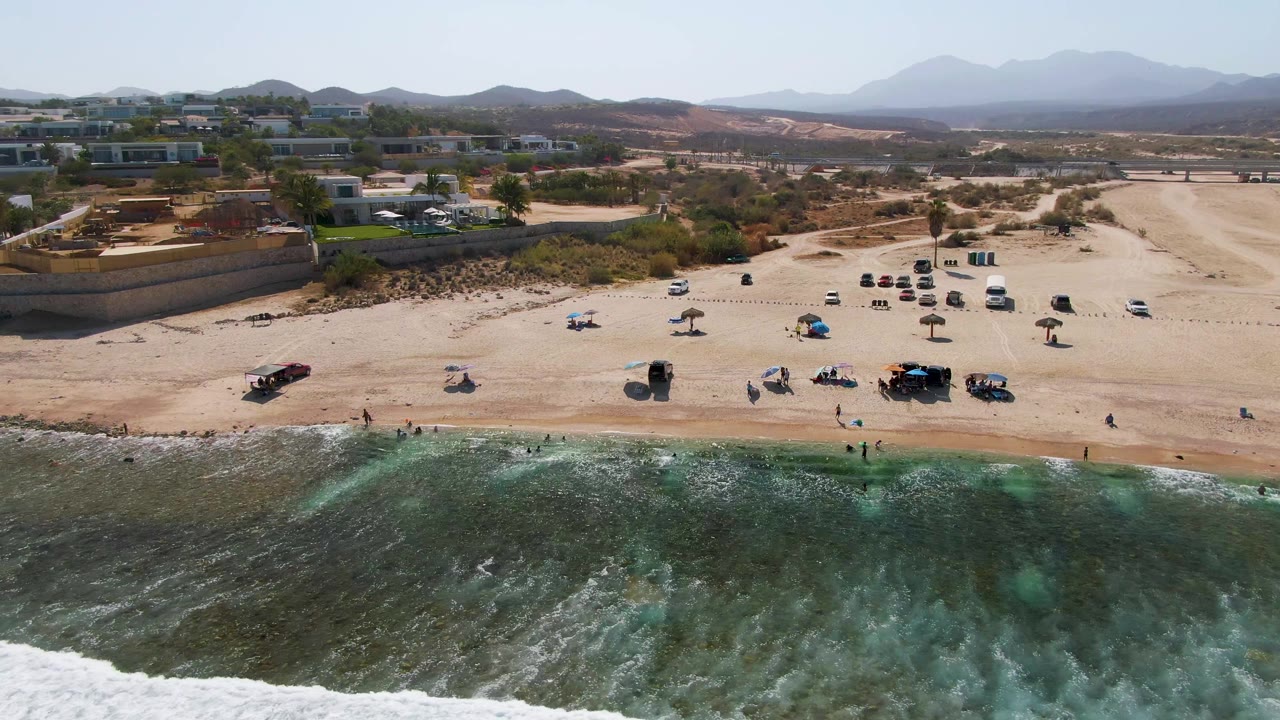 Drone view of surf, sand, beach umbrellas, parked cars, buildings, and mountains.