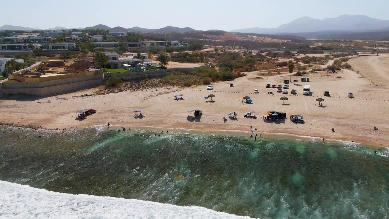 El Tule Beach aerial view with darker green water and white foam approaching the sand, people near the shoreline, parked vehicles spread across the beach, low structures inland, and pale mountains in the distance.