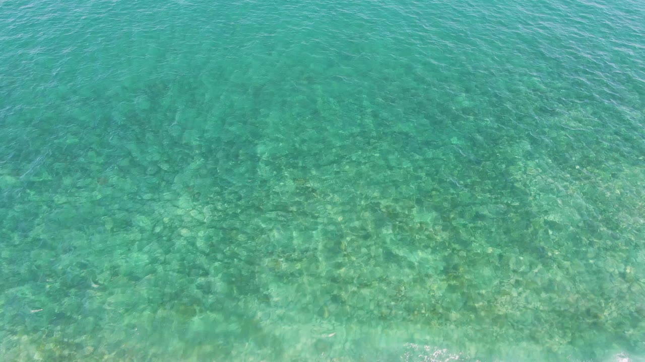 Clear blue-green water covers the frame at El Tule Beach in Los Cabos, with small ripples and mottled sand and coral texture below, no visible people.