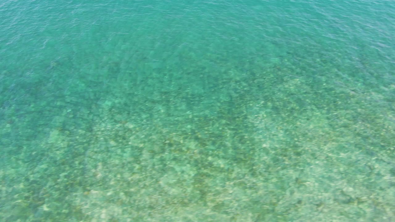 Shallow ocean water fills the frame at El Tule Beach in Los Cabos, with darker coral or rock patches visible beneath the rippled surface and no visible people.