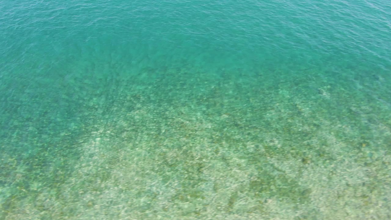 Top-down view of clear turquoise water over a pale sandy seabed at El Tule Beach in Los Cabos, with faint green patches and no visible people.