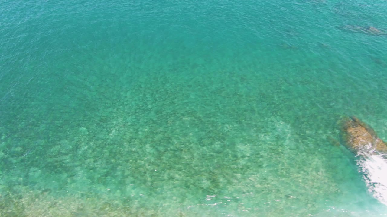 Overhead view at El Tule Beach in Los Cabos showing clear turquoise water, a tan rock outcrop near the lower center, white foam breaking around it, and no visible people.