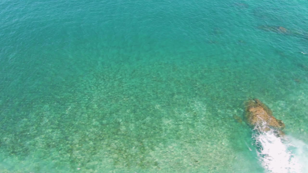 Aerial frame from El Tule Beach in Los Cabos with the main rock slightly right of center, pale seabed visible below, white surf trailing downward, and no visible people.