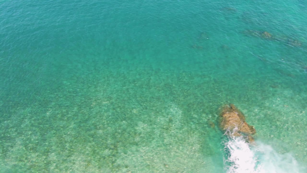 Top-down view at El Tule Beach in Los Cabos where a tan rock sits near the right edge, white foam spreads below it, the seabed remains visible, and no visible people appear.