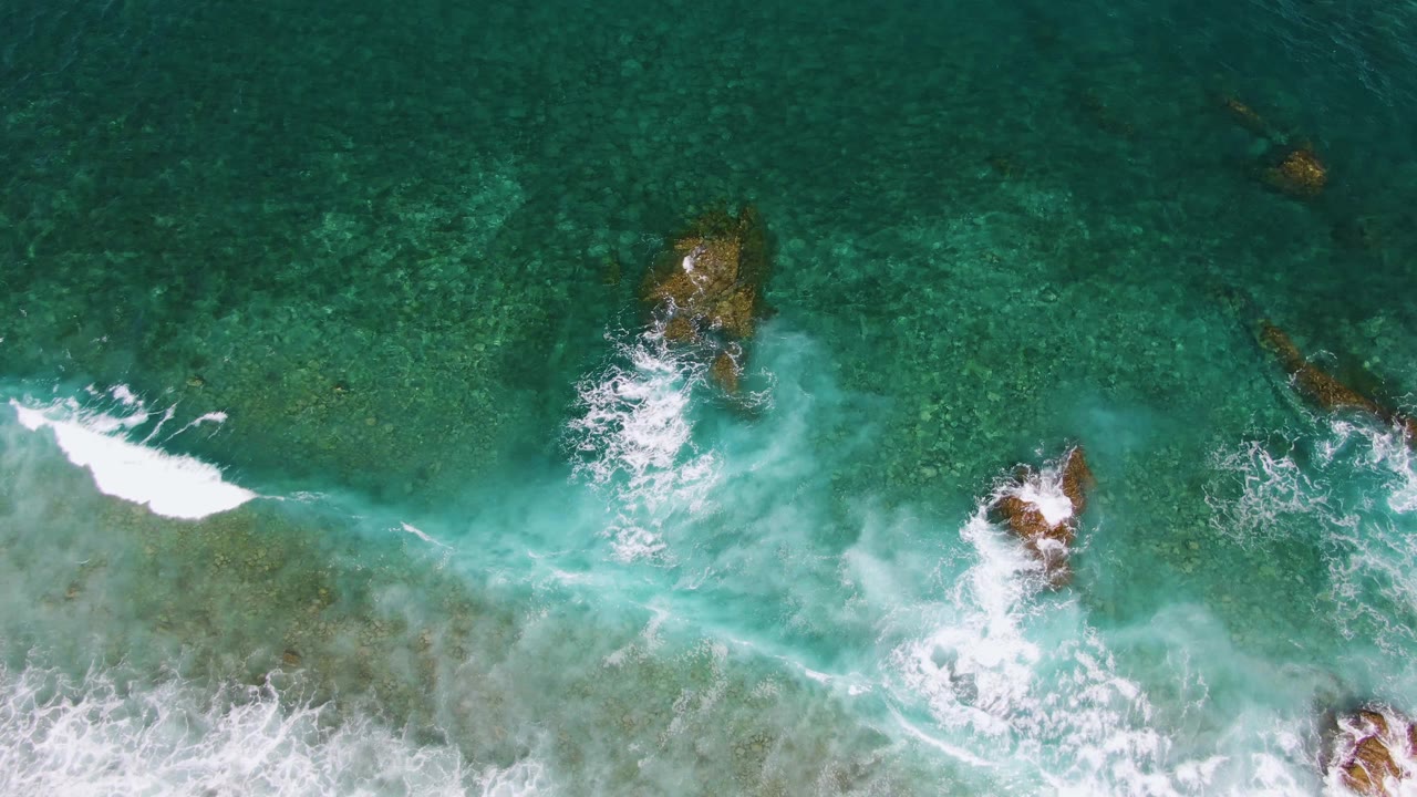 A white wave line cuts across clear green-blue water at El Tule Beach in Los Cabos, with two rocky outcrops visible below the surface and no visible people.