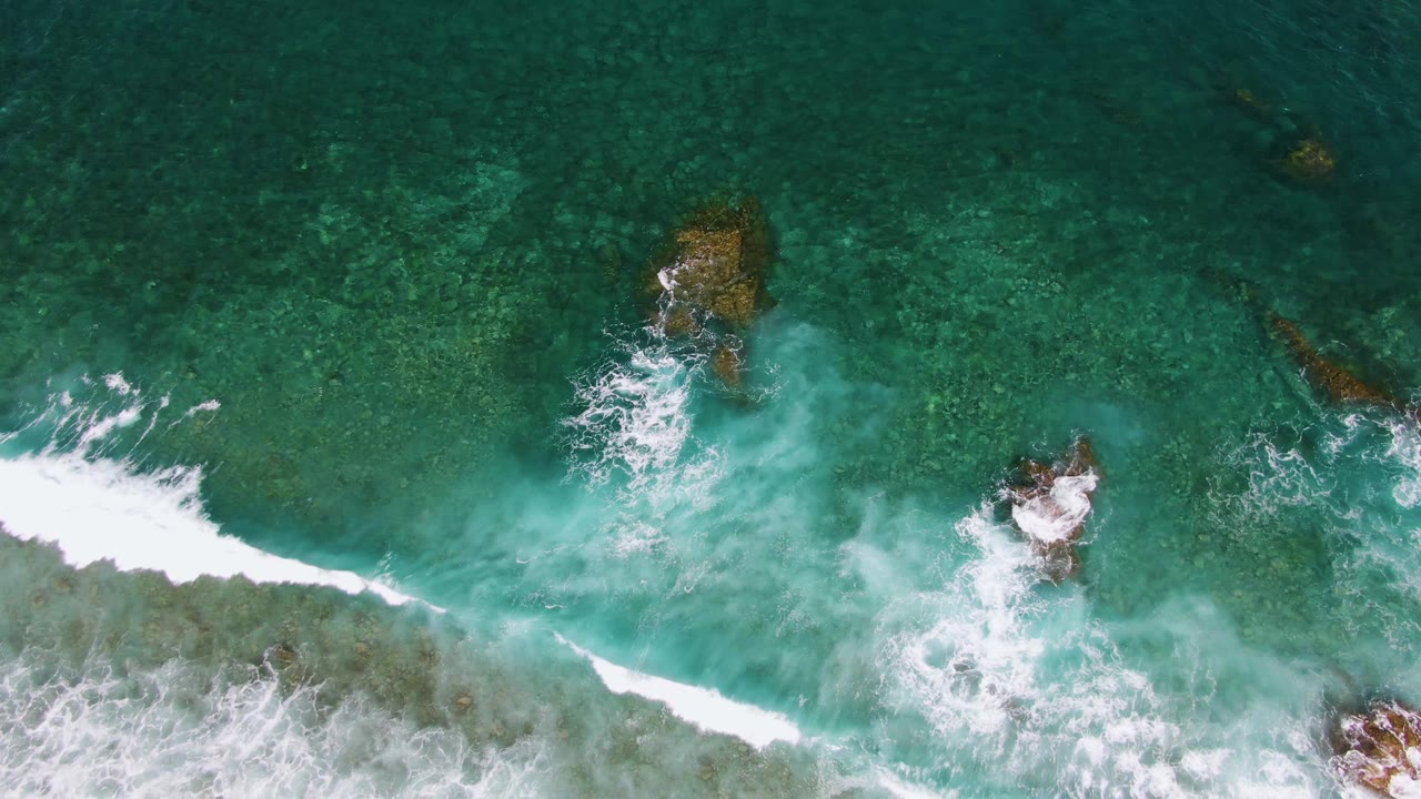 Foam spreads around several submerged rocks in clear turquoise sea at El Tule Beach in Los Cabos, with the brightest surf concentrated near the lower half and no visible people.