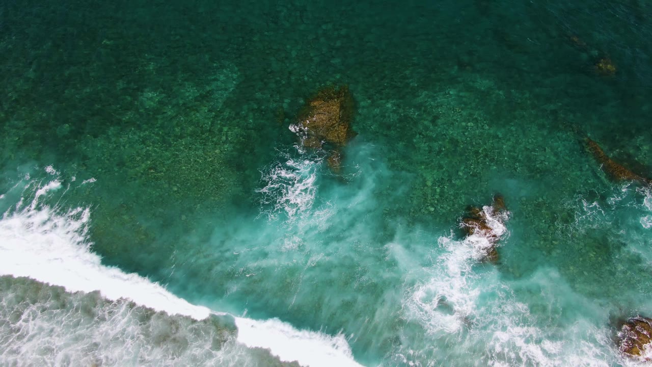 Aerial view at El Tule Beach in Los Cabos where broad white surf pushes in from the lower left across green water and exposed-looking rocks, with no visible people.
