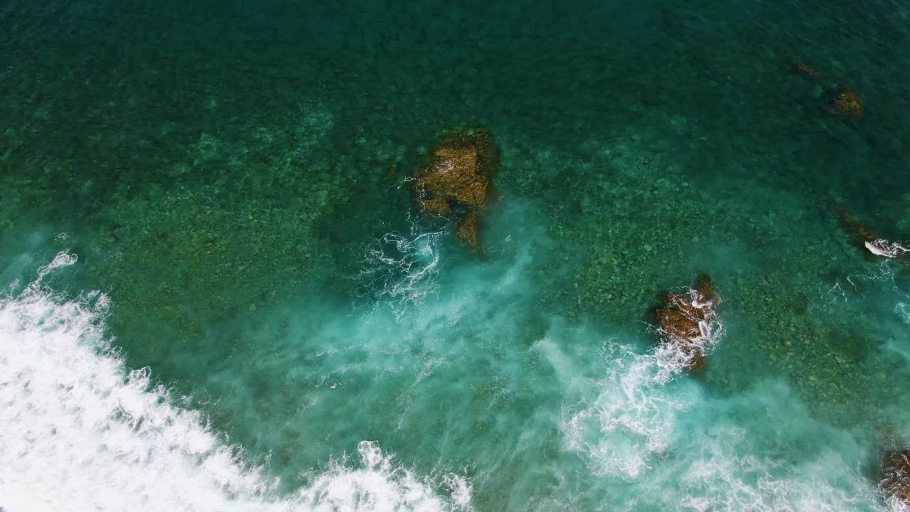 At El Tule Beach in Los Cabos, a top-down aerial frame shows white surf entering from the lower left over clear turquoise water and submerged rocks, with no visible people.