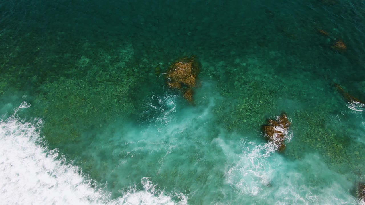 At El Tule Beach in Los Cabos, the surf has advanced farther across the lower frame while brown rock clusters sit in clear green-blue water, with no visible people.