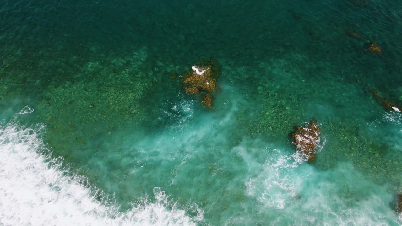 At El Tule Beach in Los Cabos, a curling band of white foam wraps around the rocks against darker green water near the top of frame, with no visible people.