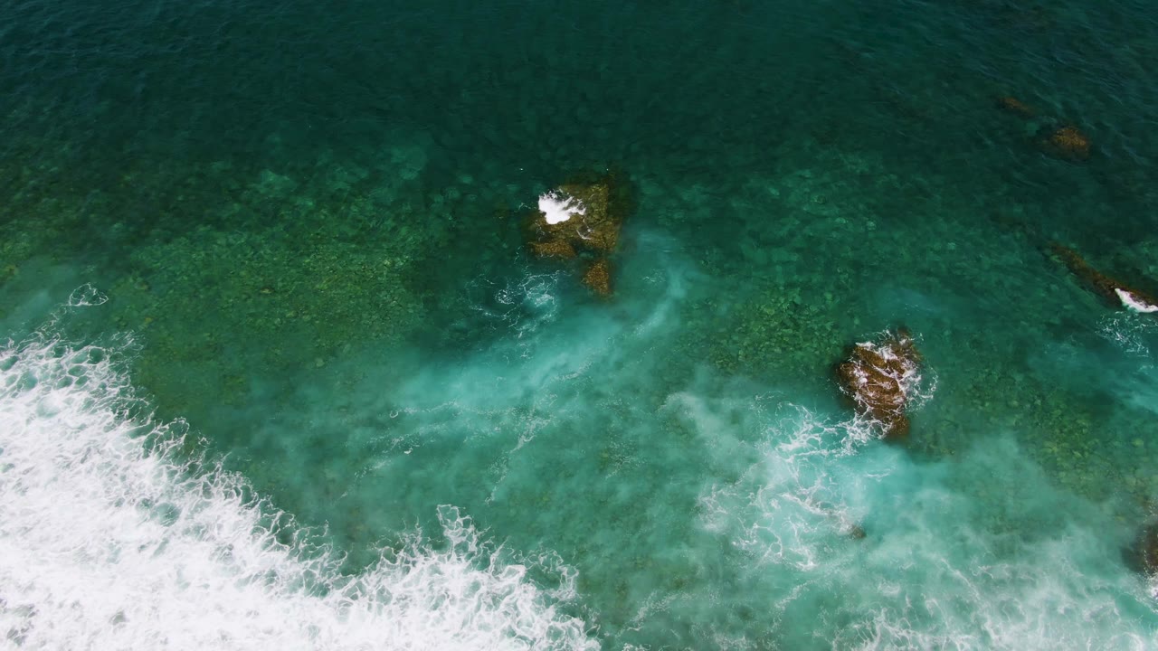 Overhead ocean frame with waves breaking from the lower left around rocks.