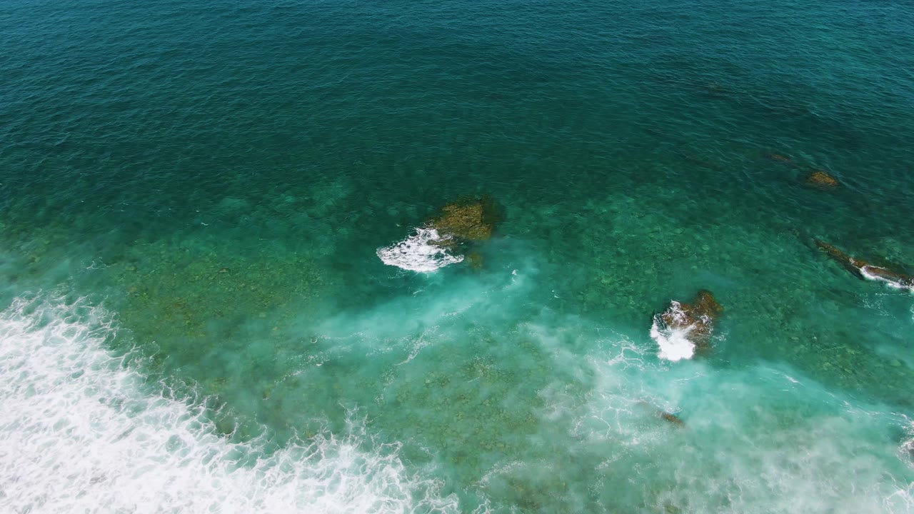 Overhead view of turquoise water and white foam breaking over shallow rocks at El Tule Beach in Los Cabos, with no visible people.