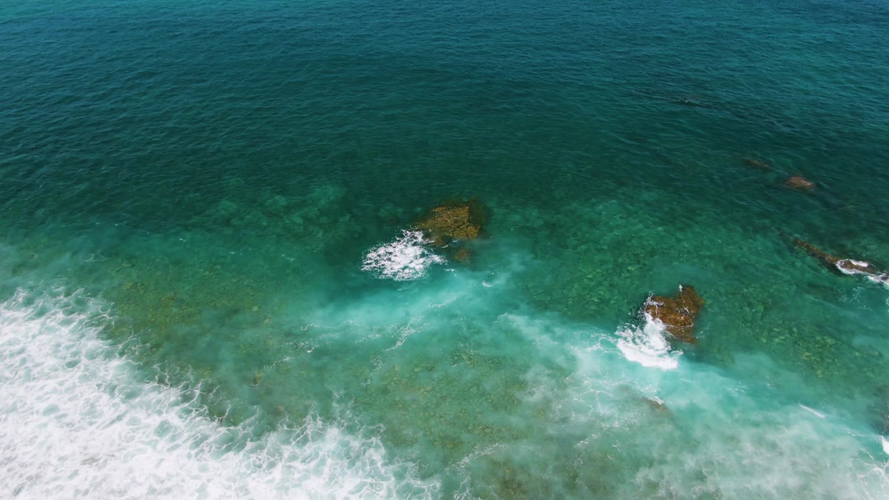 Aerial frame of white surf spreading across clear blue-green water around shallow rocks at El Tule Beach in Los Cabos, with no visible people.