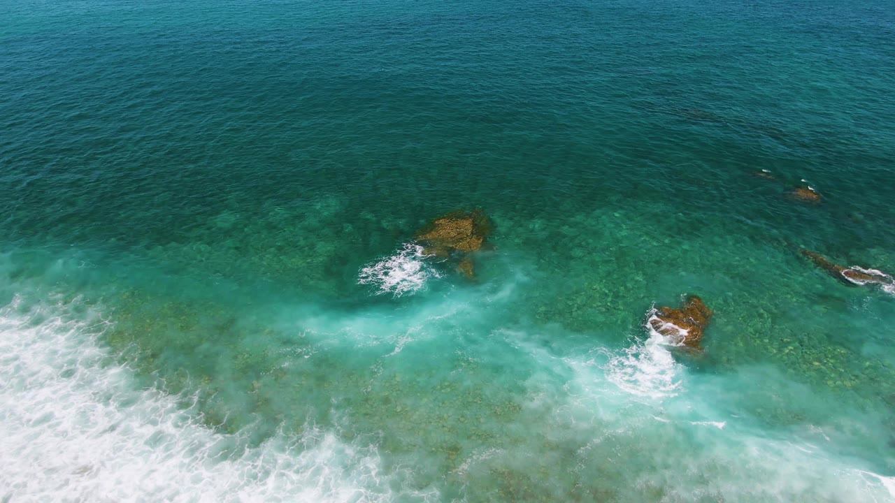 Top-down view of foam curling around two visible rock areas in blue-green ocean water at El Tule Beach in Los Cabos, with no visible people.