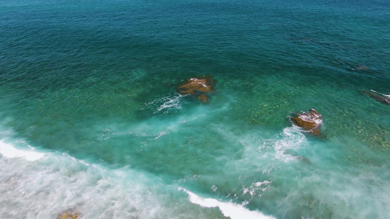 Aerial view at El Tule Beach in Los Cabos showing turquoise water, shallow rocks, white foam entering from the lower left, and no visible people.