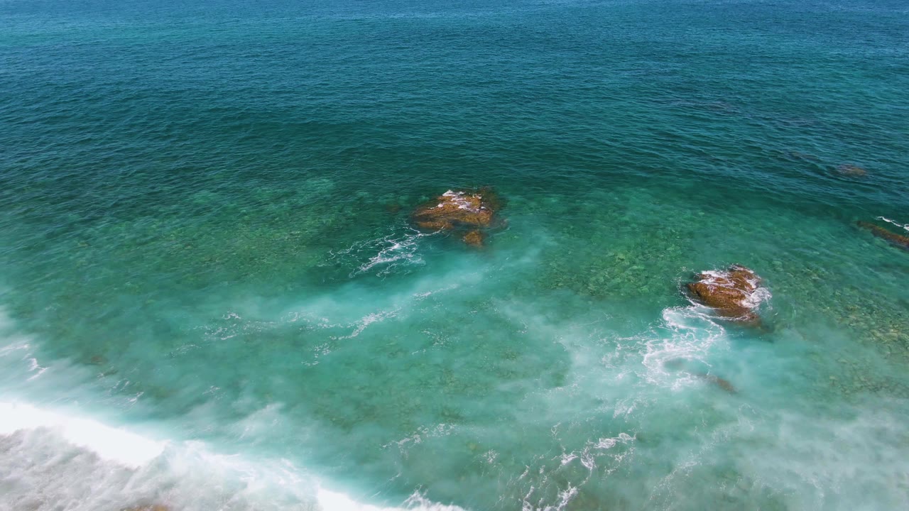 Aerial frame from El Tule Beach in Los Cabos with foamy surf spreading through clear turquoise water around two brown rocks and no visible people.