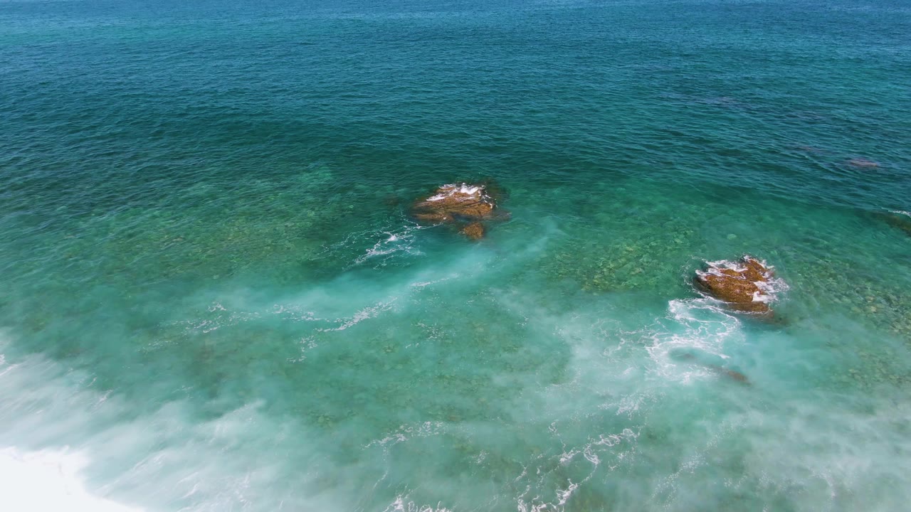 Aerial view at El Tule Beach in Los Cabos showing blue-green water, visible submerged rocks, a foamy wave line near the bottom edge, and no visible people.