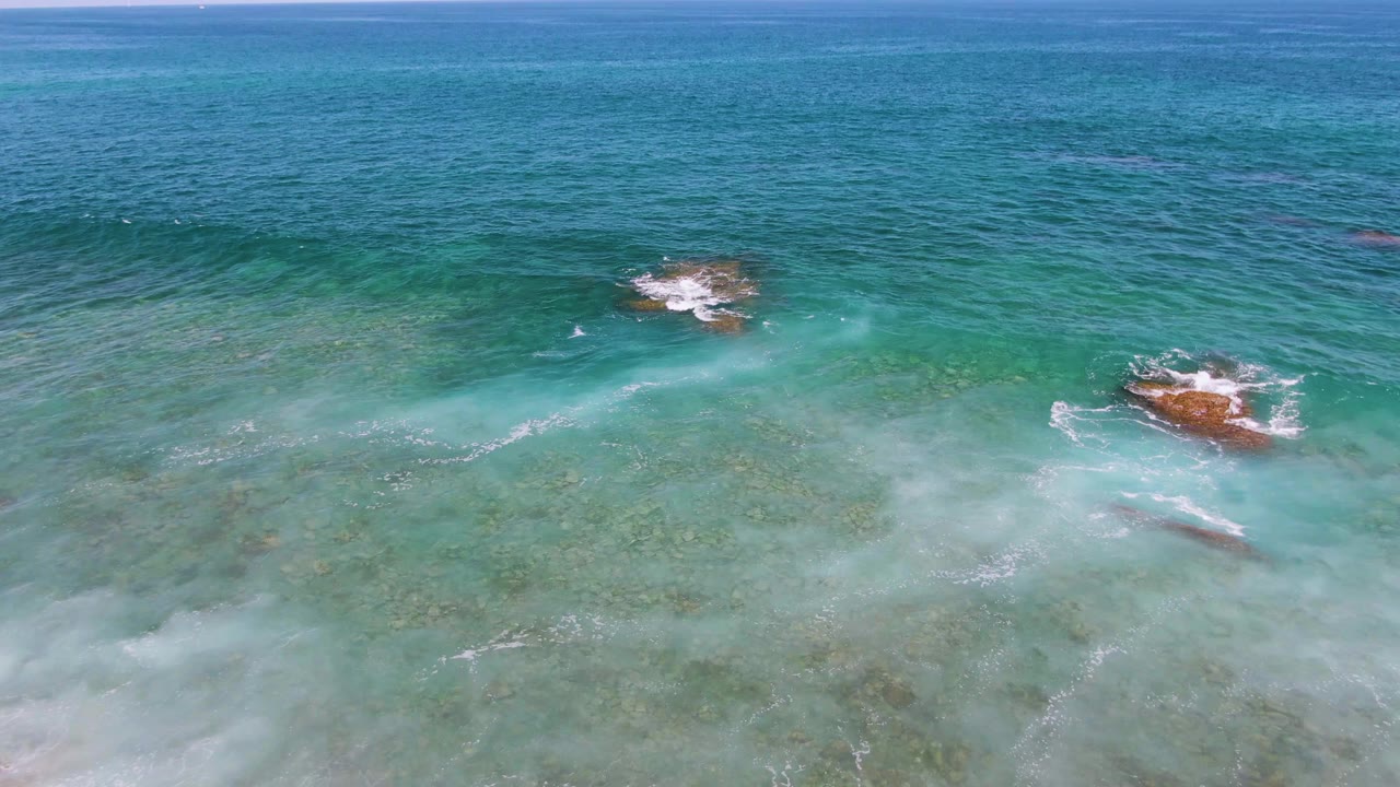 Aerial view of turquoise shallows at El Tule Beach in Los Cabos, with white foam forming around shallow rocks and no visible people.