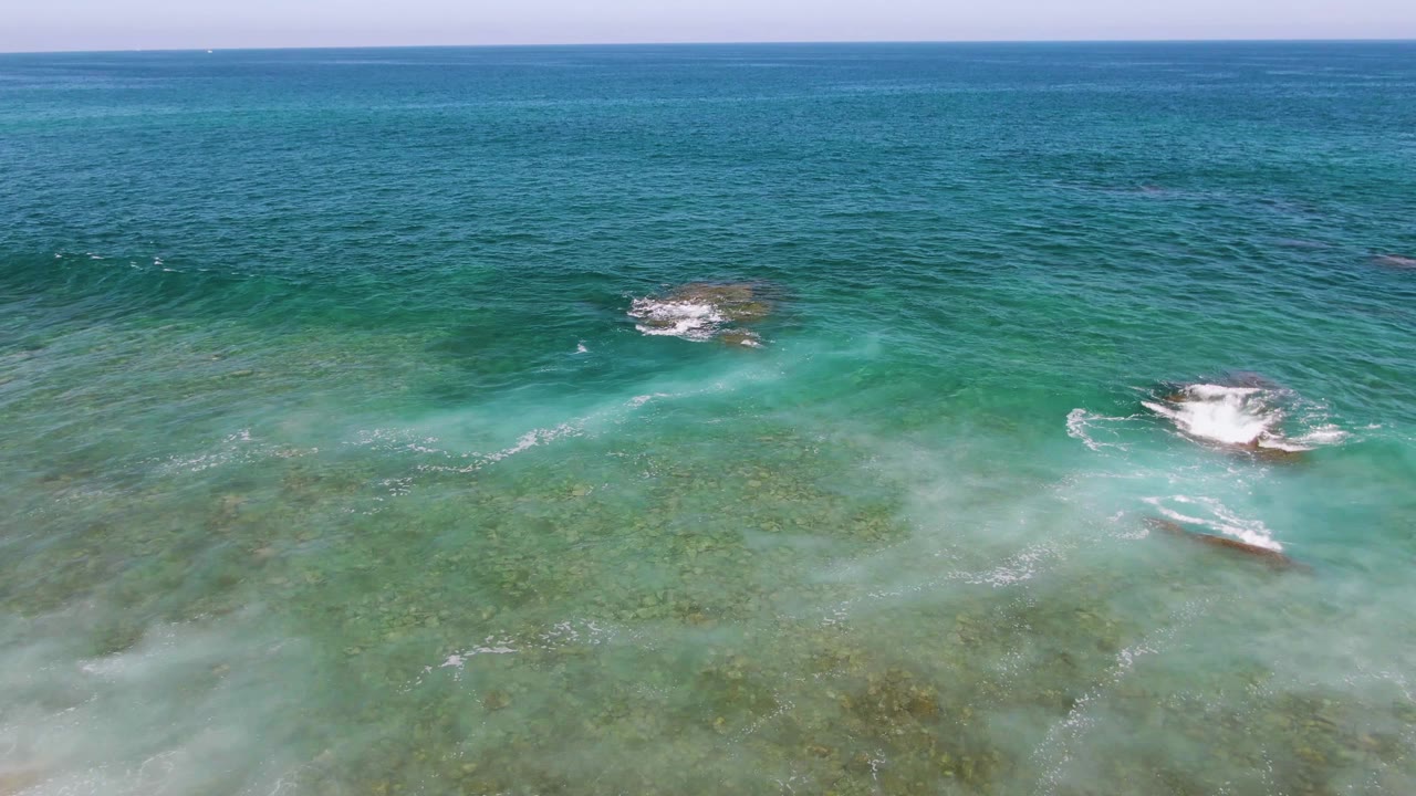 Aerial frame from El Tule Beach in Los Cabos showing foam sliding over a shallow rock while darker blue ocean sits beyond the turquoise water and no visible people appear.