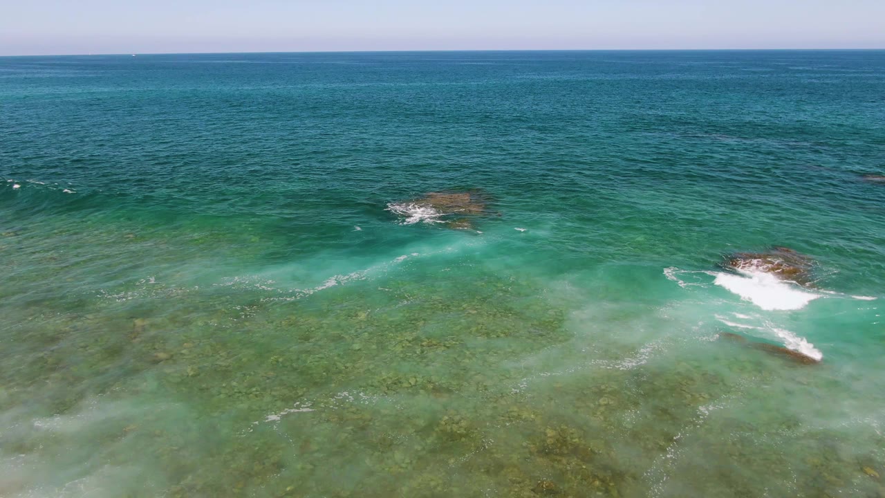 Aerial view at El Tule Beach in Los Cabos with a breaking wave on the right, submerged rock patches in green water, open blue sea in back, and no visible people.