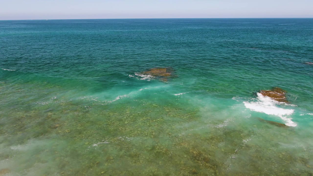 Aerial frame from El Tule Beach in Los Cabos showing a low band of white surf moving across shallow green water, with scattered rocks and no visible people.