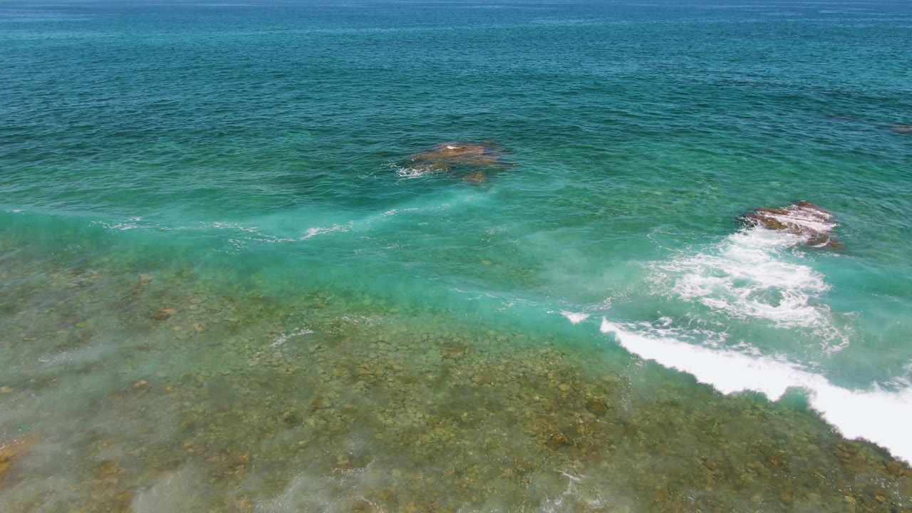 Aerial frame from El Tule Beach Los Cabos showing turquoise water, submerged rock texture, white surf pushing across the lower frame, and no visible people.