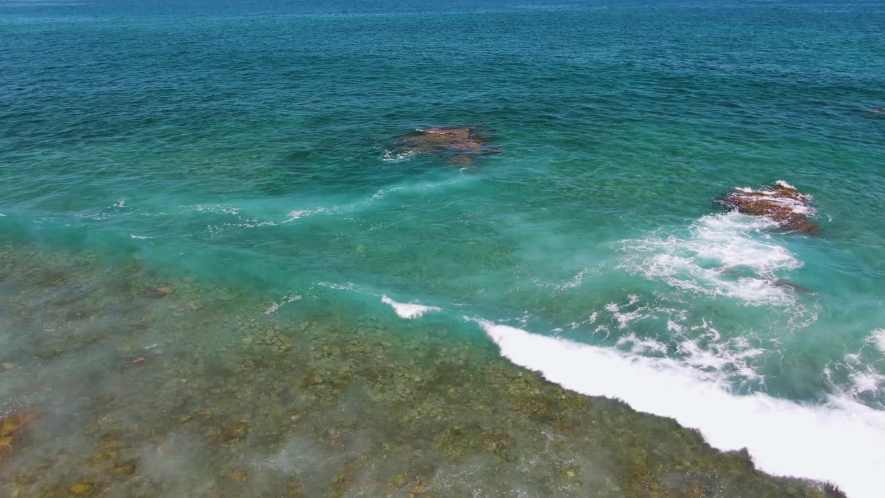 Aerial view at El Tule Beach Los Cabos with a broader band of white surf crossing the foreground, rocky outcrops in the middle distance, and no visible people.