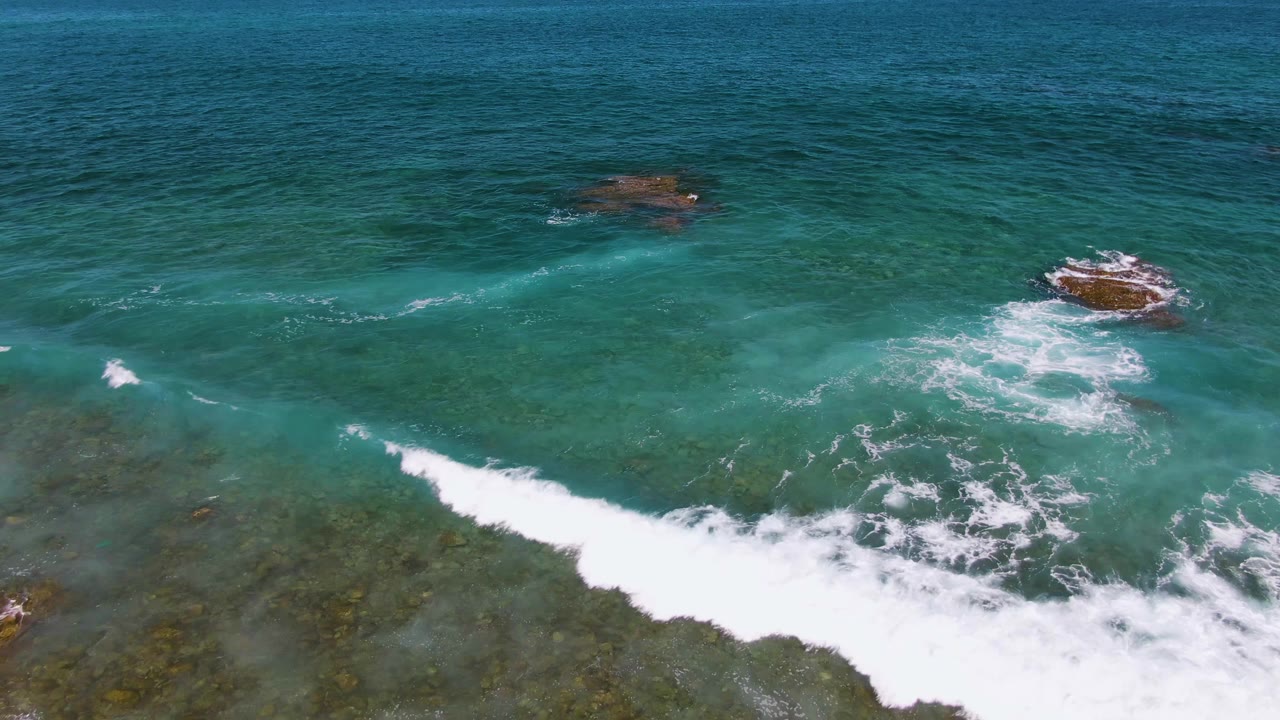 Aerial frame from El Tule Beach Los Cabos showing white water curling around a central rock, darker blue open water above it, and no visible people.
