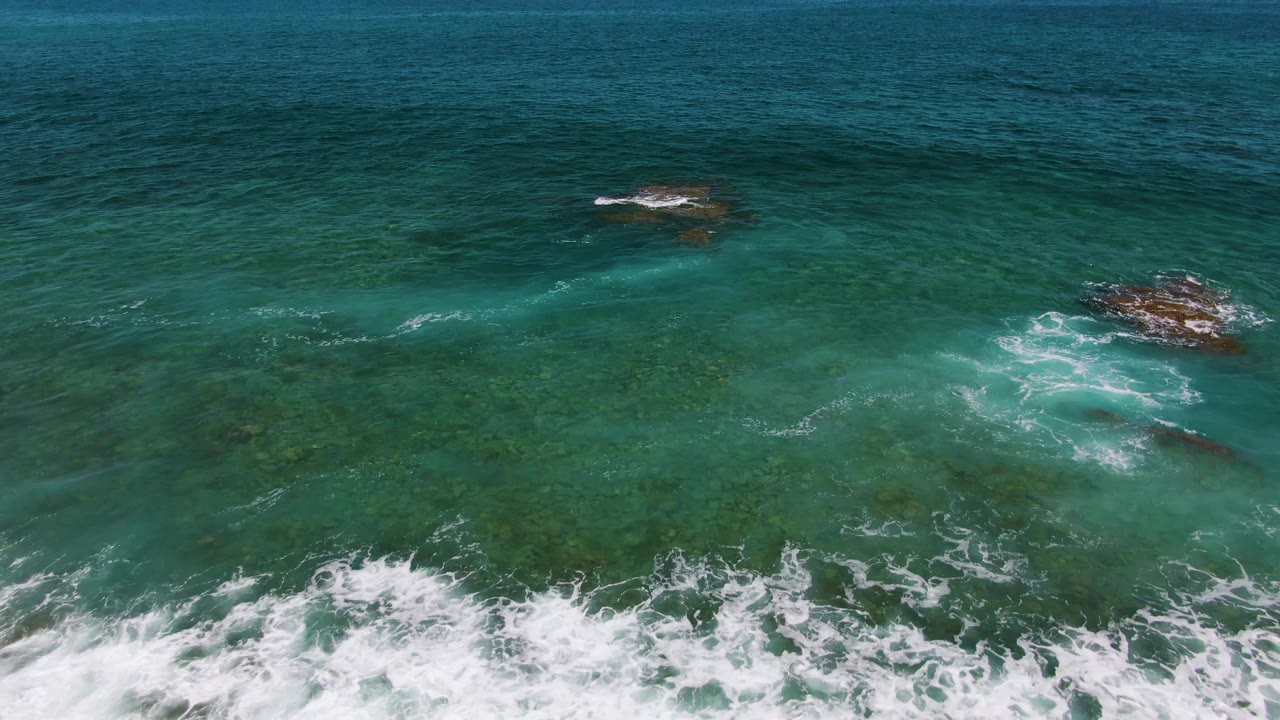 Aerial view of turquoise water at El Tule Beach in Los Cabos with white foam crossing shallow rocks near the lower edge and no visible people.