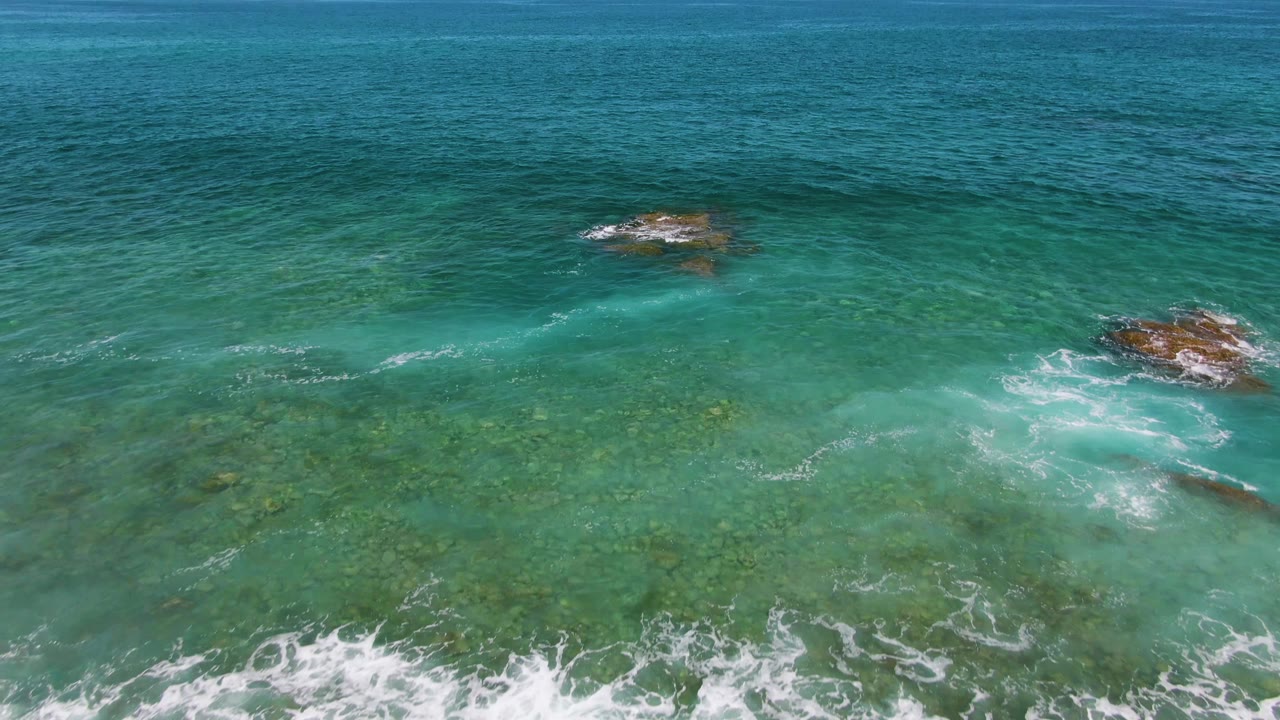 Aerial view at El Tule Beach in Los Cabos with clear green-blue water, scattered rock outcrops, thinning foam, and no visible people.