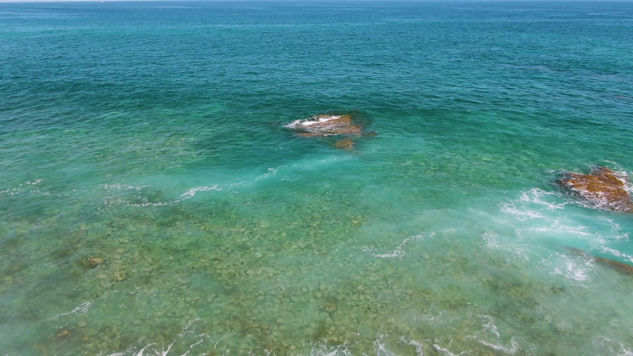Aerial frame from El Tule Beach in Los Cabos showing shallow turquoise water, a rocky patch at upper right, faint surf trails, and no visible people.