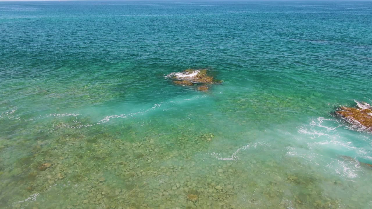 Aerial view at El Tule Beach in Los Cabos with a wider expanse of clear water, exposed rock patches, pale foam near the lower right, and no visible people.
