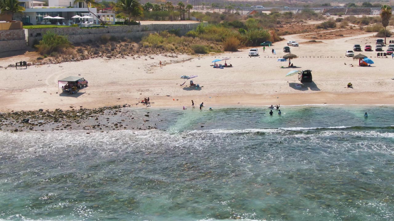Aerial frame over El Tule Beach Los Cabos showing white surf in the foreground, beachgoers near the waterline, and scattered umbrellas and vehicles on the sandy shore.