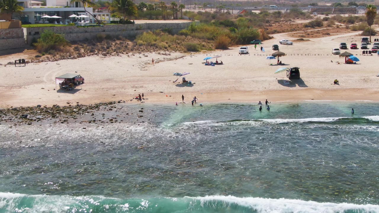 Beachgoers stand in shallow water as small waves approach a sandy shore.