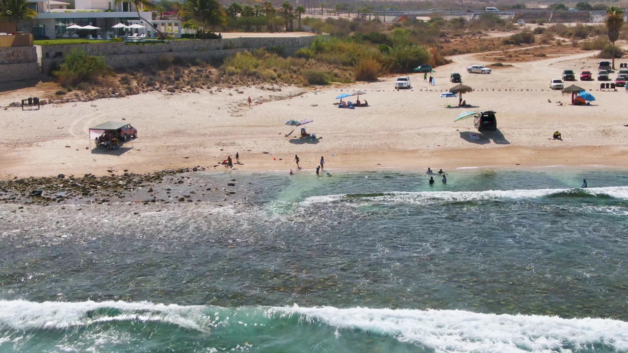 El Tule Beach Los Cabos seen from above as a broader white wave breaks in the foreground and small figures move along the waterline near umbrellas and parked vehicles.