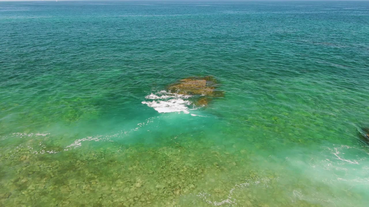 At El Tule Beach in Los Cabos, the rock remains near center while a thin white wave line drifts through the shallow green-blue water and no visible people appear.