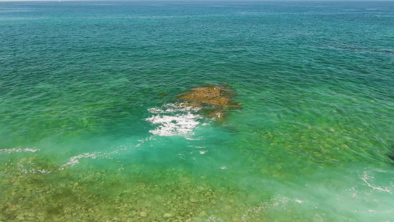 At El Tule Beach in Los Cabos, white foam collects on the lower-left side of the rocky outcrop as clear water reveals the seabed and no visible people are present.