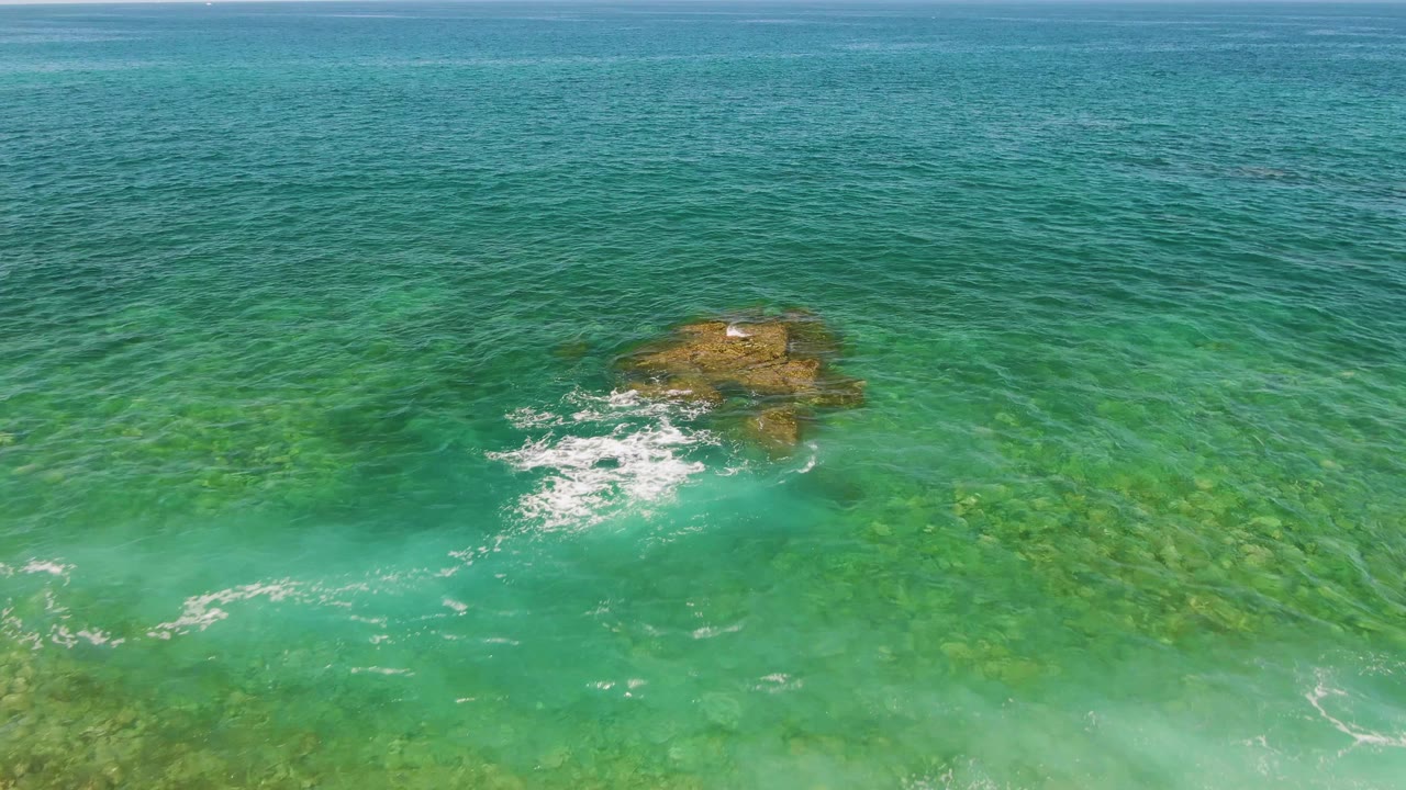 At El Tule Beach in Los Cabos, the rocky outcrop sits lower in frame with white foam trailing below it, green seabed texture visible through the water, and no visible people.